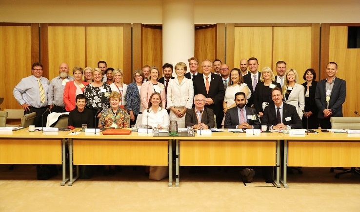 Photo of a group of men and women in business attire behind six people seated at desks. The Prime Minister and Minister are standing in the middle.