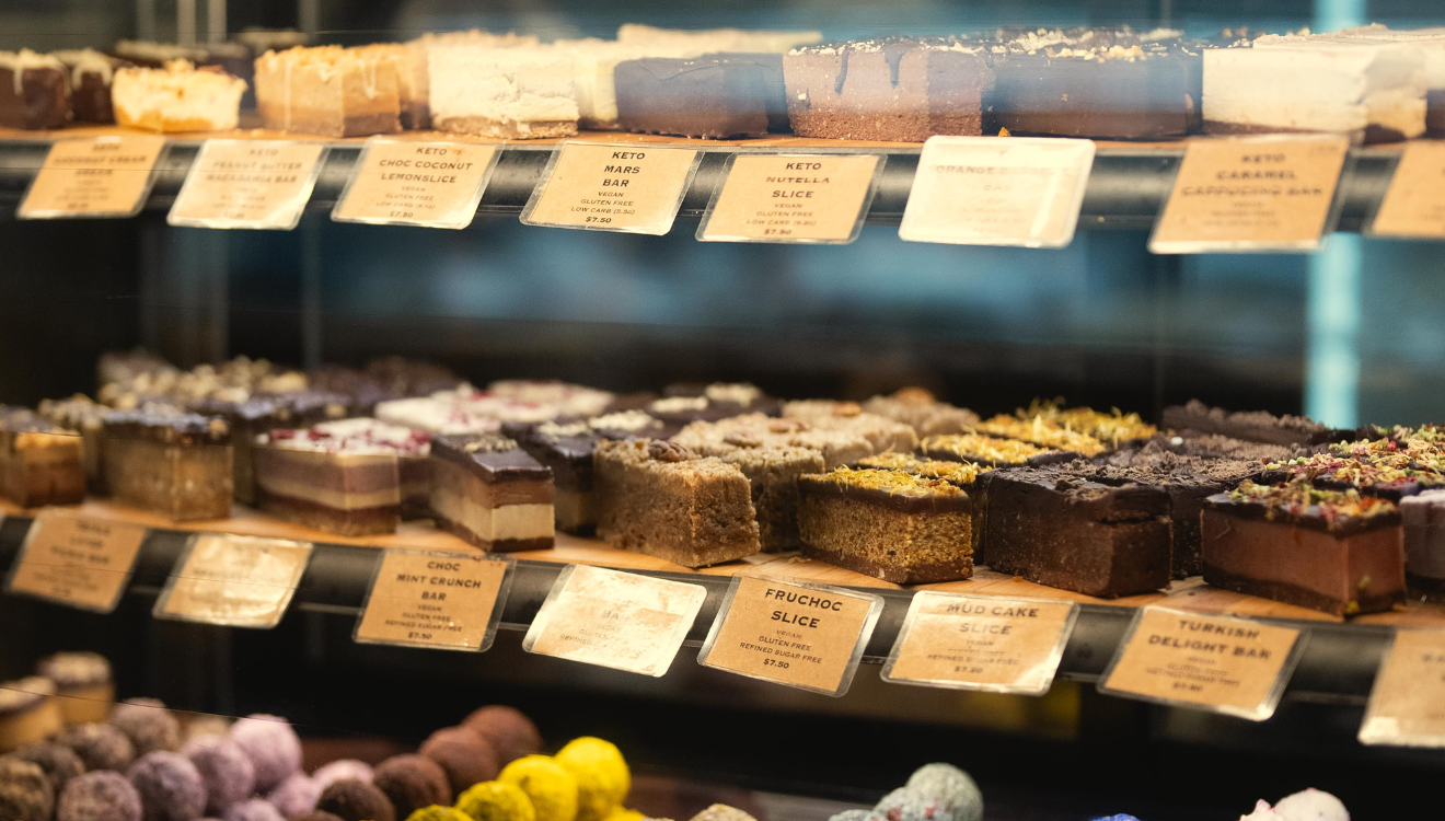 Three shelves behind a glass counter displaying various bakery items with price labels.