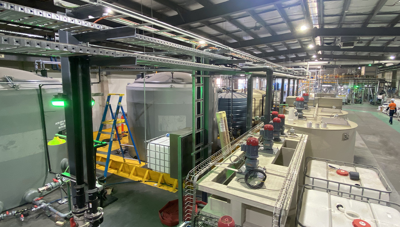 An industrial processing facility showing large storage tanks, overhead piping, pumps, control equipment, and maintenance ladders. 