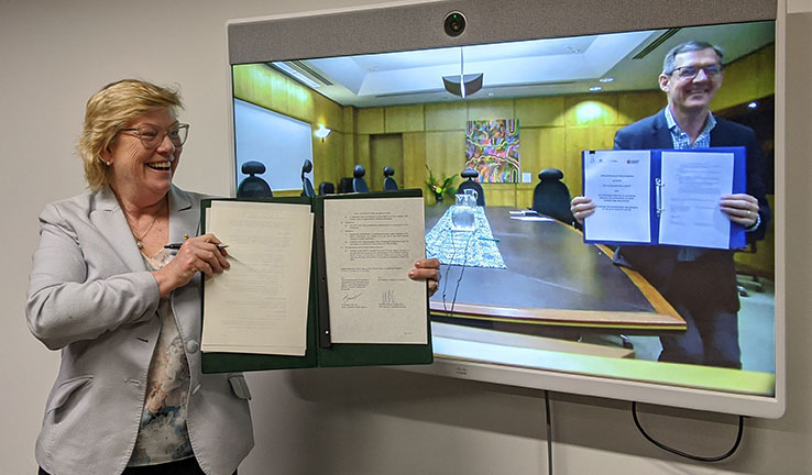 Photo of a woman standing in front of a TV screen with a man on the screen, both holding up documents