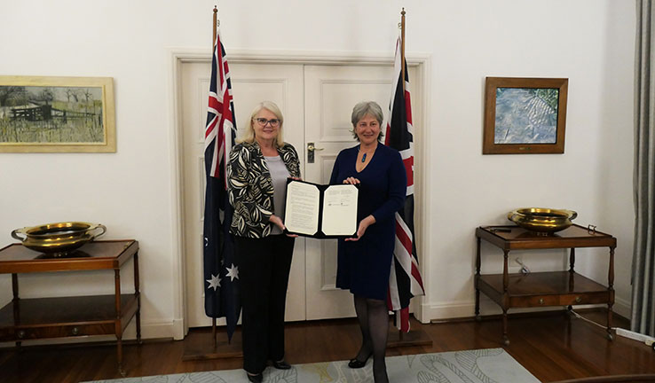2 women standing in from of a door, holding a document between them and with the Australian and UK flags behind them