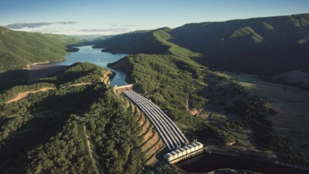 Snowy Hydro Tumut 3 Power Station. Aerial view of Snowy Hydro Limited Tumut 3 hydroelectric Power Station, with multiple pipes leading down from a dam amidst green landscape.