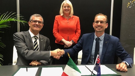 Two men sitting at a table shaking hands, with a woman standing behind them