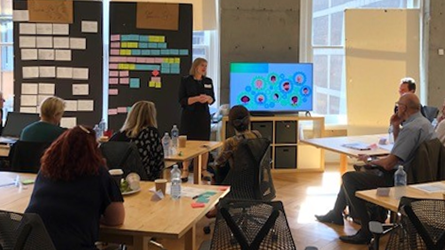 The Director addresses the workshop attendees A woman talking to a group of adults sitting at desks. A dark board with small pieces of paper pinned to it is behind her