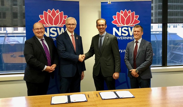 Four men standing behind a table, the middle two shaking hands.