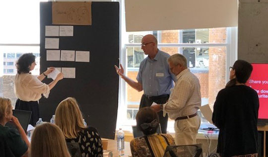 A woman pinning pieces of paper on a dark board. Beside the board, three attendees wait their turn to share ideas.