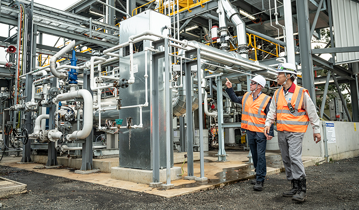 Photograph shows engineers from Australia and Japan wearing reflective safety vests, safety hard hats and safety glasses walking around the hydrogen plant. There are pipes and equipment in the background.