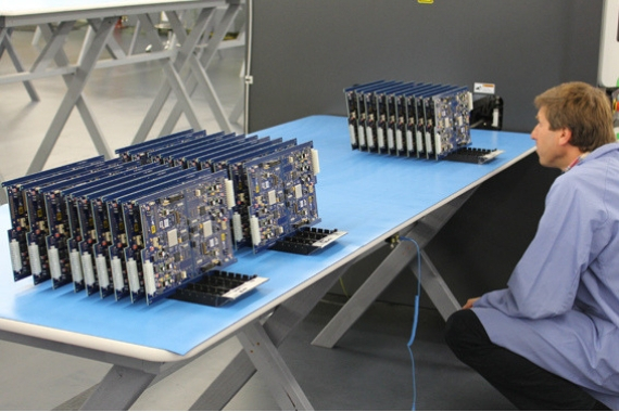 Multiple blue circuit boards manufactured by Puzzle Precision sitting on top of a clean blue table being inspected by a crouching man.
