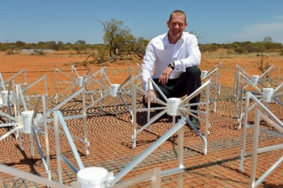 Professor Steven Tingay crouches within a tile of knee-high, spider-like antennas, which form part of the Murchison Widefield Array, surrounded by red dirt, sparse vegetation and blue sky.
