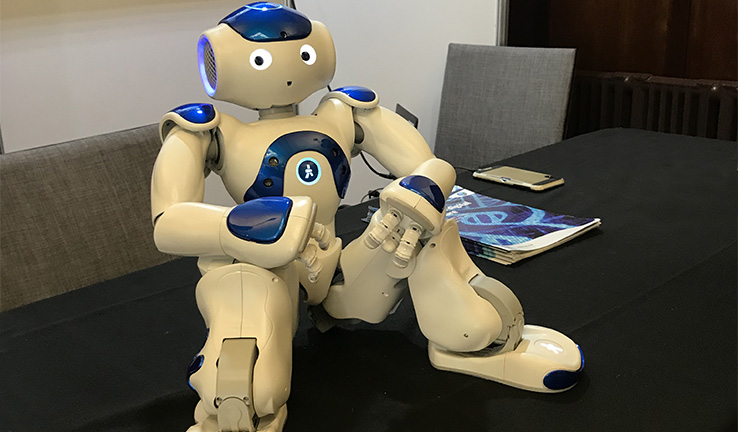 Photo of a robot that looks like a friendly human sitting on a table at the exhibition. Photo of a robot that looks like a friendly human sitting on a table at the exhibition.
