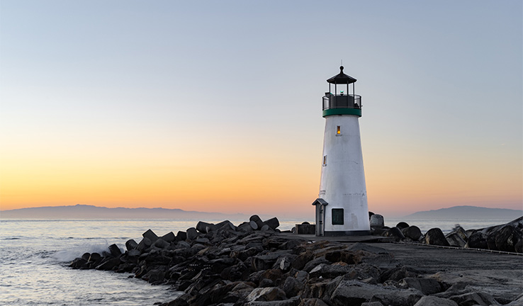 Photo of a lighthouse on a rocky outcrop by the sea at sunset. Photo of a lighthouse on a rocky outcrop by the sea at sunset.