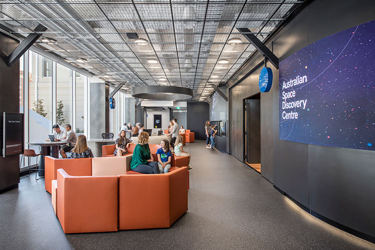 A room full of orange couches with people sitting on them looking relaxed with a poster of the 'Australian Space Discovery Centre' behind them.