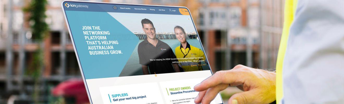 Business man holds a laptop and browses for information on the ICN Gateway website. He’s standing outside on a grass verge in front of a building.