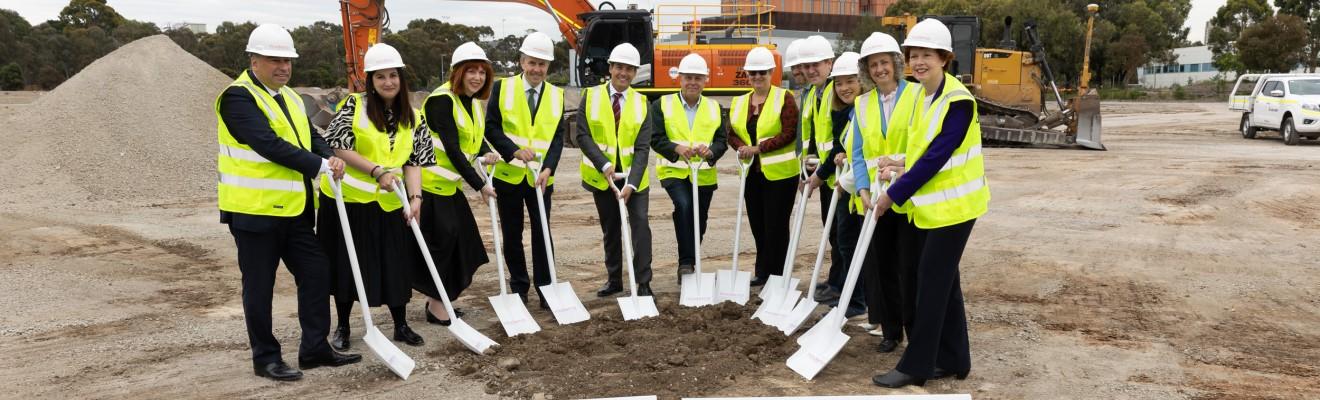 Twelve men and women in hard hats and high-vis vests stand in a semi-circle holding shovels on a building site. 