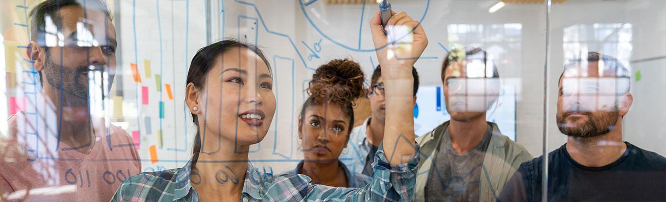 A diverse group of business people watch a female colleague draw business growth projections on a clear window.