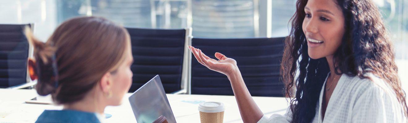 Two businesswomen have a conversation at a table with their laptops and coffee.