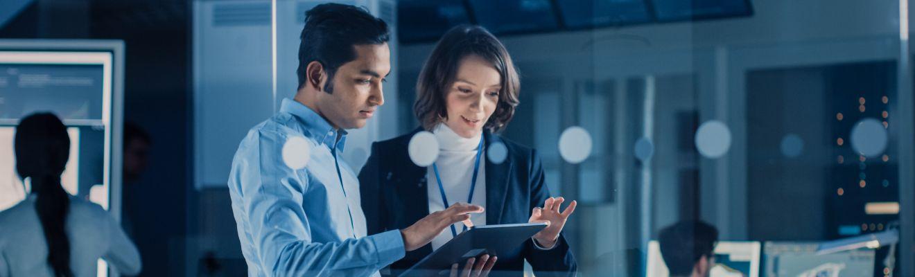 Two co-workers are standing together and discussing what they see on a computer tablet. In the background there are 2 other people working on computers.
