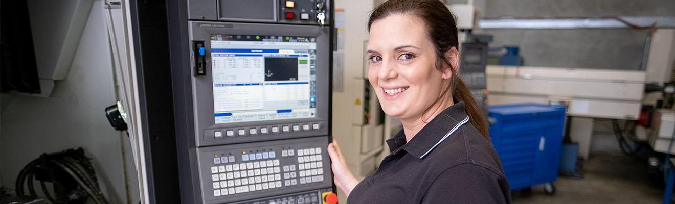 Image shows a woman in a factory. She is standing next to the control panel for a machine tool, smiling and looking towards the camera.