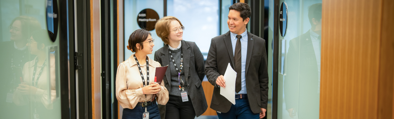 Three staff from diverse backgrounds walk down a corridor looking at each other smiling. 