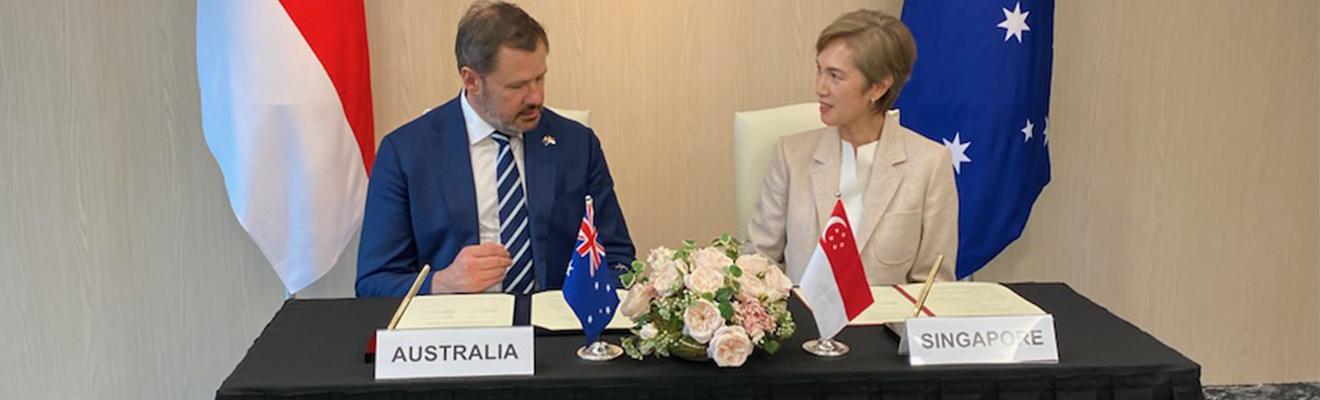 Minister Husic and Minister Teo, sitting at a desk signing the MoU. They are surrounded by flags of Australia and Singapore. 