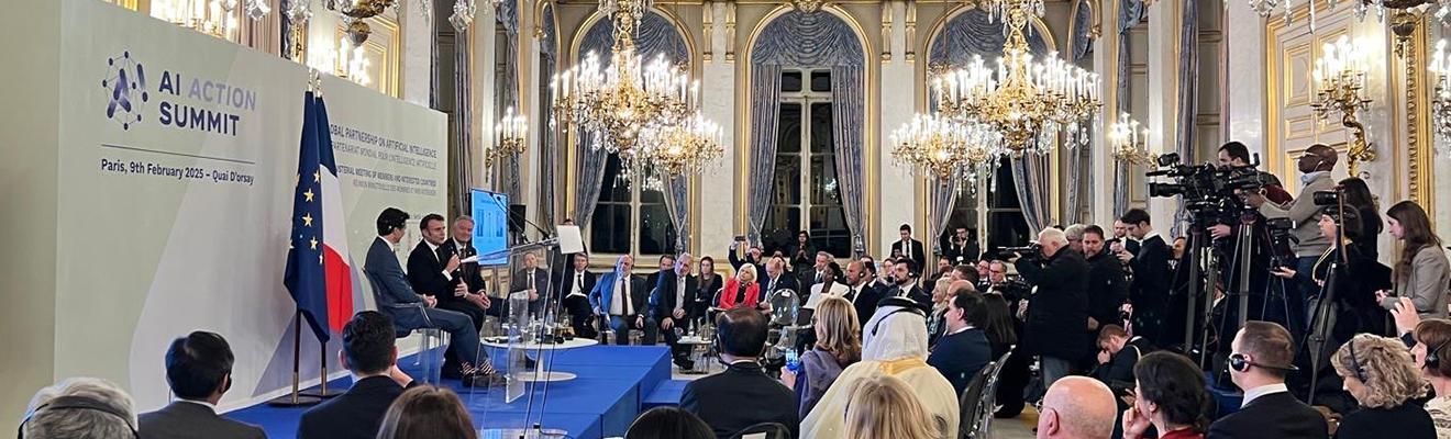 Photo of the AI Action Summit stage located in the Grand Palais. Several political figures are sitting down and talking to a room filled with people. 