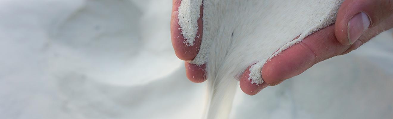A pair of hands is picking up white silica sand and letting it fall through their fingers