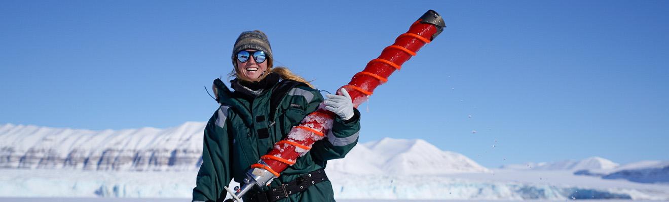 Female polar marine biologist stands in a snowy landscape holding an oversized drill bit