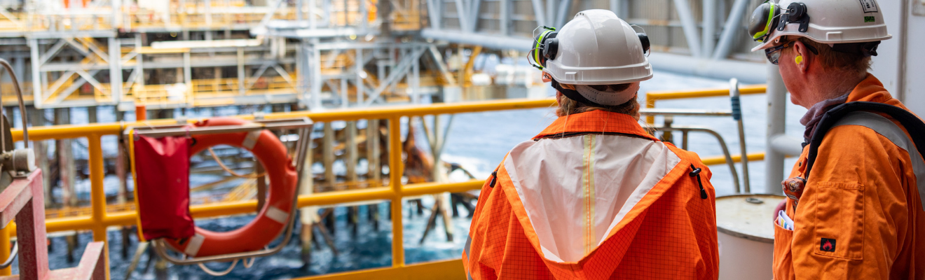 Two oil and gas workers wearing hard hats and orange gear look out from the back of a vessel onto an offshore rig.