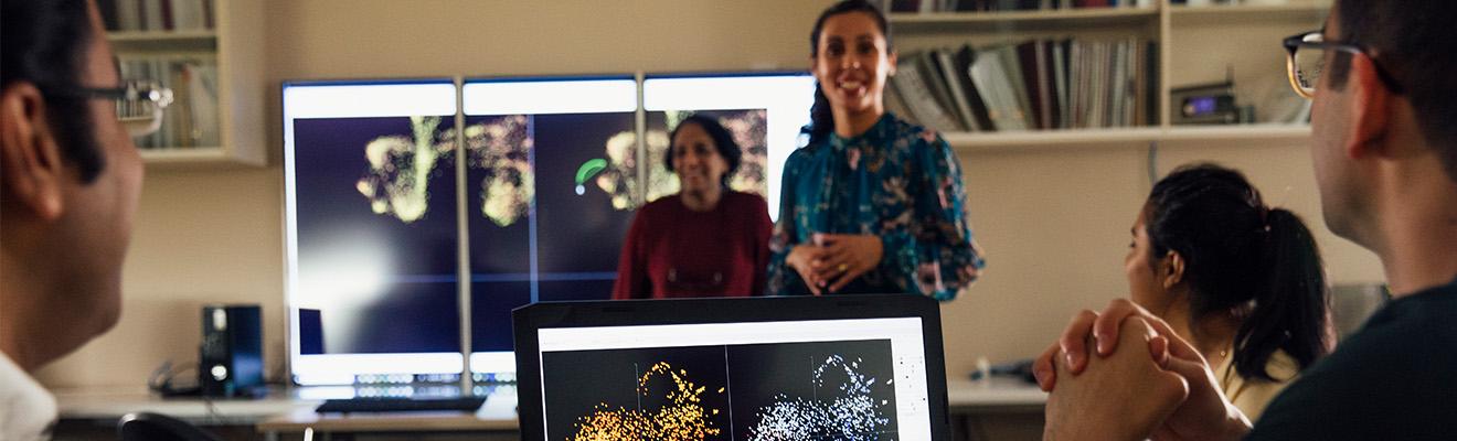 A group of people watching colleagues present on a technology topic. They're watching screens with data plot displays.