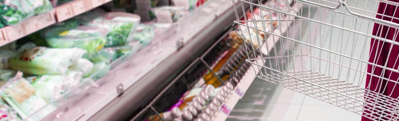 Person with shopping basket in front of salad and juice items in a supermarket aisle