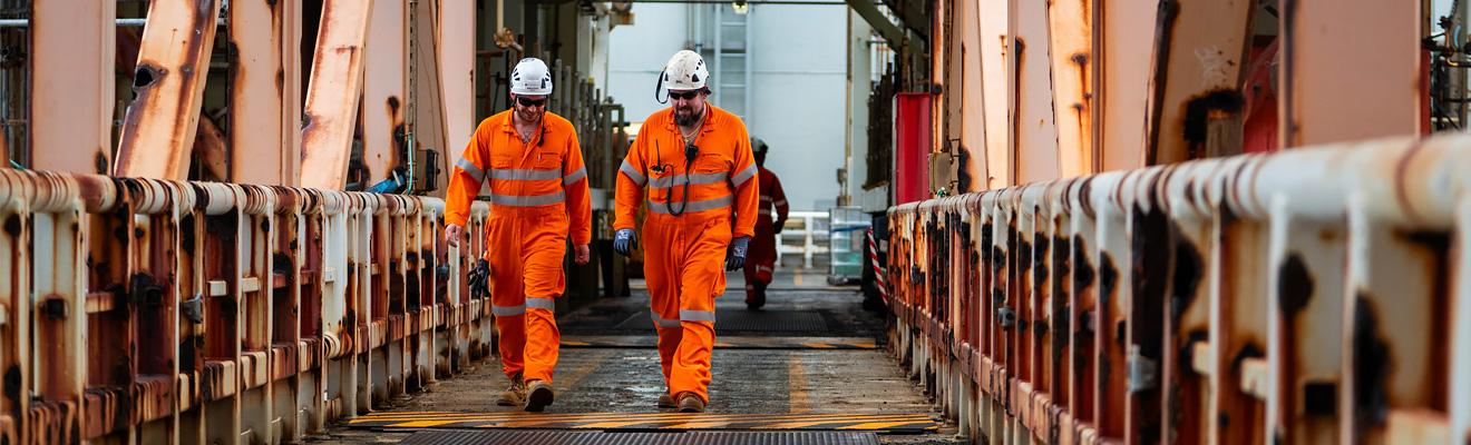 Two crew members in high visibility overalls and hard hats walk down a rusty walkway