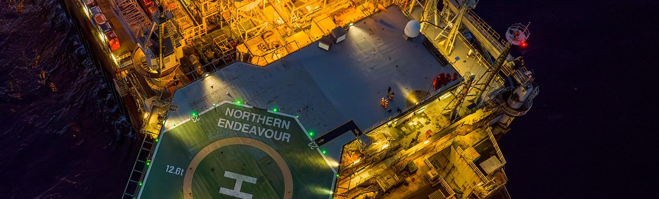 An aerial view looking down at the Northern Endeavour at night. The topsides and helideck are lit up.