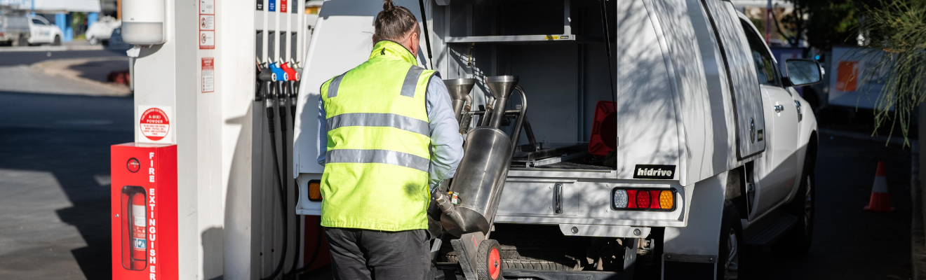 A field officer wearing a high visibility vest unloads fuel testing equipment from the back of a utility vehicle at a service station.