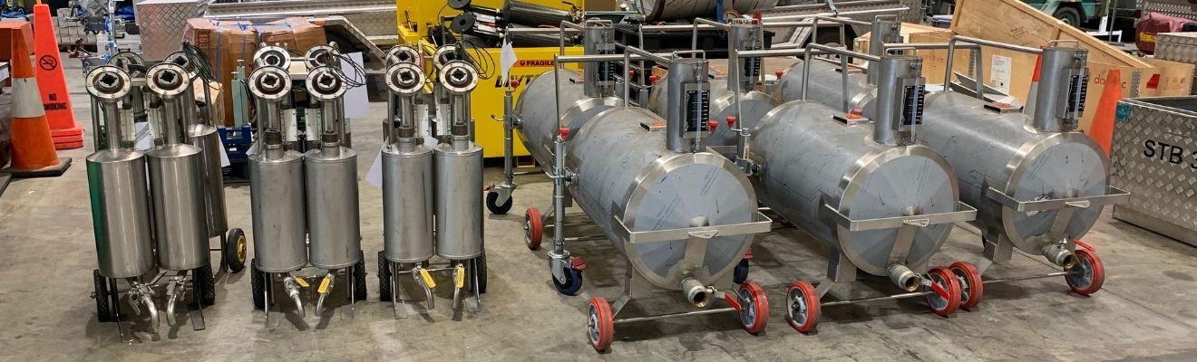 Stainless steel fuel measurement equipment on wheels, including several cylindrical tanks and smaller measurement units, arranged inside a warehouse ready for transport or deployment.