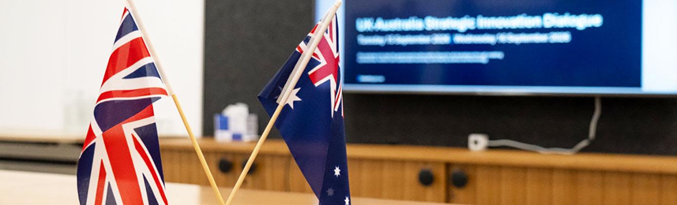British and Australian flags on a conference room desk with a presentation screen in the background ‘UK-Australia Strategic Innovation Dialogue’. 