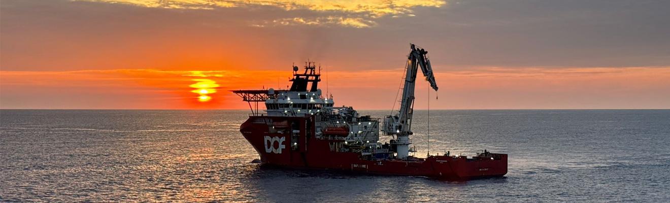 A red support vessel with the crane extended sits in a calm sea with a blazing orange sunset in the background