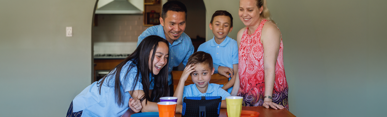 Two parents and their 3 children are smiling while they stand and sit around their dining table as they talk to family overseas using a tablet.