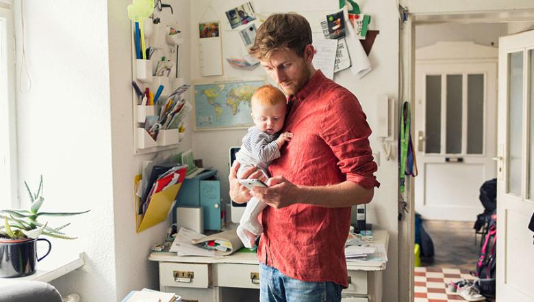 A young man working in his home office holds a baby while checking his phone