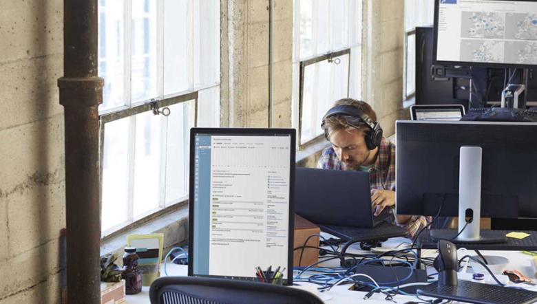 A man with headphones leans over his laptop, concentrating on his work. He’s in an informal office with desks and large computer screens