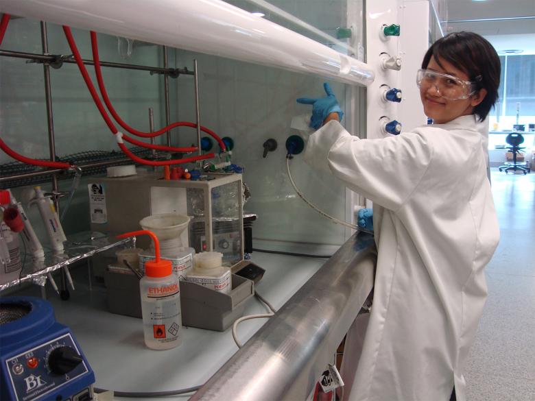 Photo of Tich-Lam Nguyen standing in front of a lab bench with various pieces of scientific equipment. She is wearing a lab coat, lab goggles and latex gloves. 