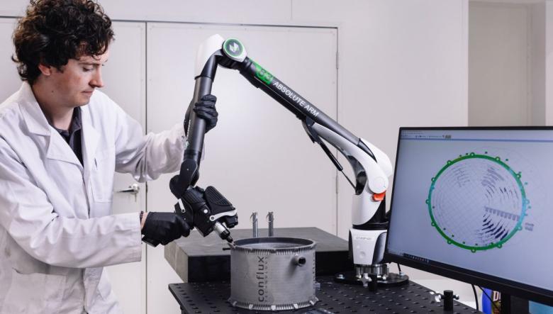 A person in a white lab coat holds a laser scanning arm above a 3D printed heat exchanger. A computer monitor displays a virtual 3D model.