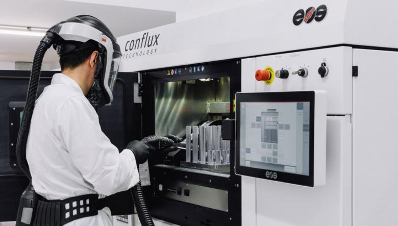 A person stands in front of an open 3D printer door and is wearing protective clothing to protect from fumes.