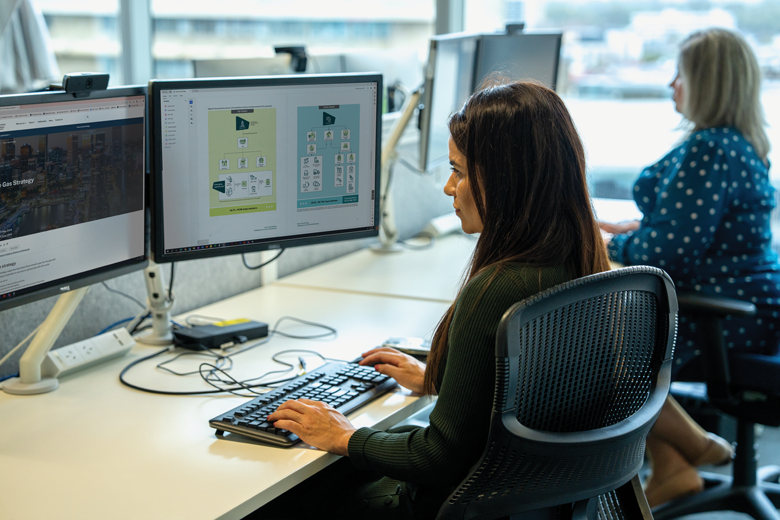 Two middle-aged white women sitting at their desks working in a well-lit office. The person in the foreground is typing on a keyboard, focused on a monitor displaying diagrams and charts. In the background, another person is engaged at a similar workstation.  