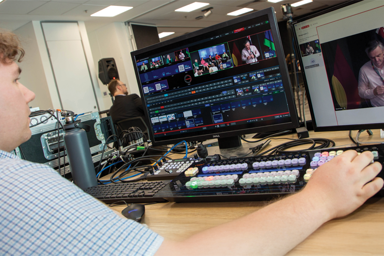 A younger man seated at a desk with multiple monitors and a keyboard with coloured keys. The main screen shows video editing software with clips and timelines.