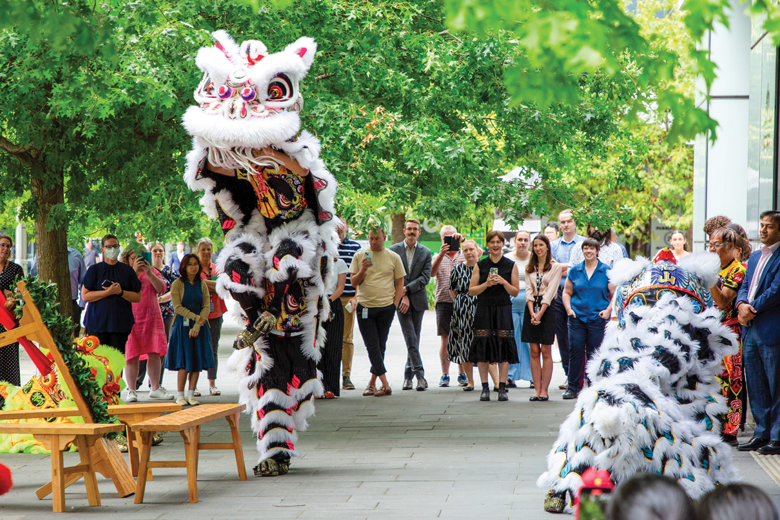 Outdoor traditional lion dance performance with 2 performers in white costumes accented with black and red, one upright, one crouching. A crowd watches nearby.