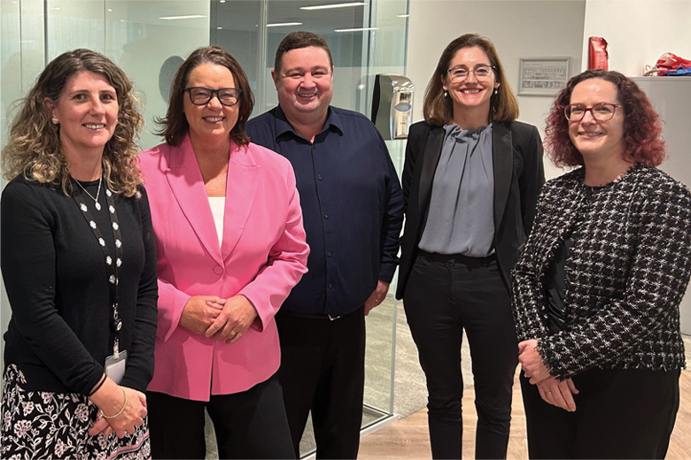 Five smiling middle-aged people (4 women and one man) standing in a line in an office.  