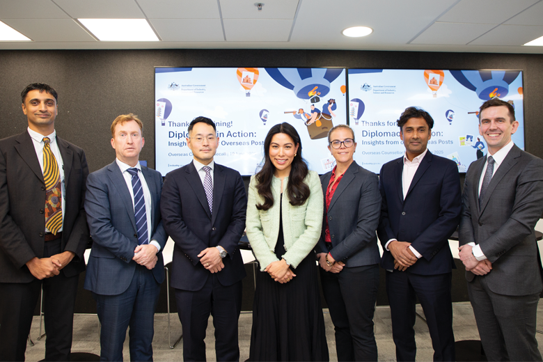 There are 7 individuals of different ethnicity (5 men and 2 female) in formal business attire standing in a row in front of 2 large screens. The screens display a slide titled 'Thanks for joining! Diplomacy in Action: Insights from our Overseas Posts'.