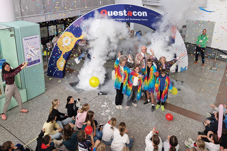 A group of people (adults and school-aged children) in colourful tie-dye shirts standing under an archway labelled 'Questacon – The National Science and Technology Centre'. Fog surrounds them, with confetti in the air and yellow balloons on the ground.