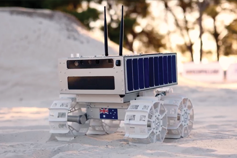 A small white 6-wheeled robotic rover on sandy terrain. It has a rectangular body with side solar panels and 2 antennas on top. An Australian flag sticker is visible on the front.