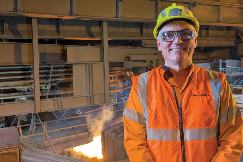 A white middle-aged man stands inside an industrial factory wearing an orange high-visibility jacket, white gloves and a yellow hard hat. Behind him is a large piece of industrial machinery with metal beams and structures. Bright light from molten material glows beneath the equipment, indicating active manufacturing processes.